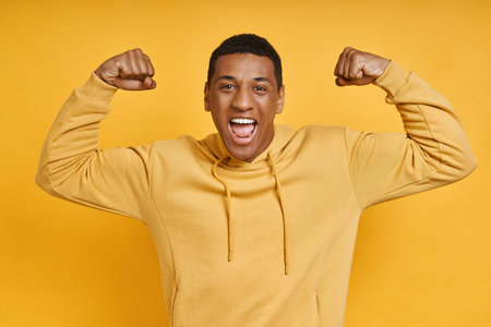 Playful mixed race man showing his biceps while standing against yellow backgroundの写真素材