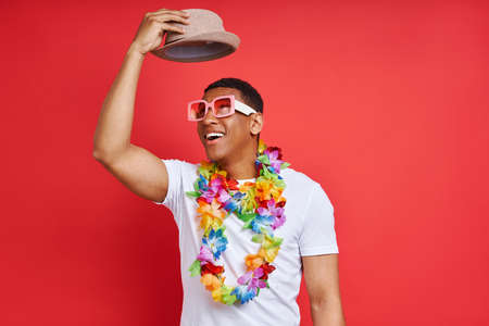 Happy young man in Hawaiian necklace carrying hat while standing against red backgroundの写真素材