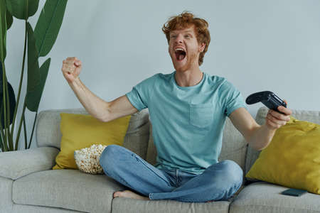 Excited redhead man playing video games and gesturing while sitting on the couch at homeの写真素材