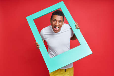 Playful mixed race man looking through a picture frame while standing against red backgroundの写真素材