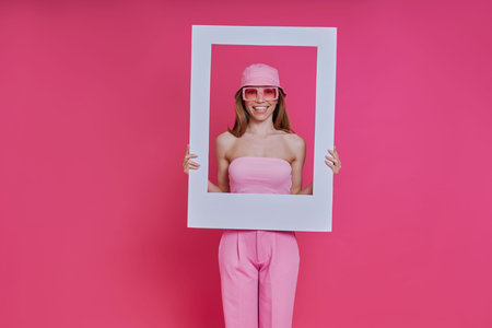 Cheerful young woman looking through a picture frame while standing against pink background