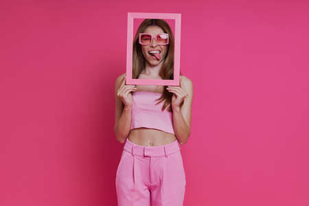 Playful young woman looking through a picture frame while standing against pink backgroundの写真素材