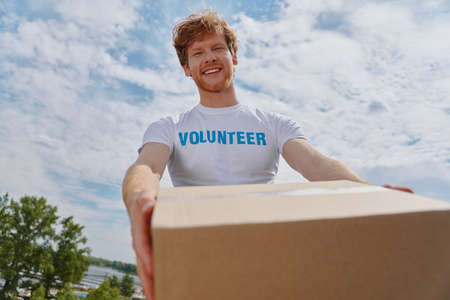 Happy young man in volunteer shirt stretching out donation box and smilingの写真素材