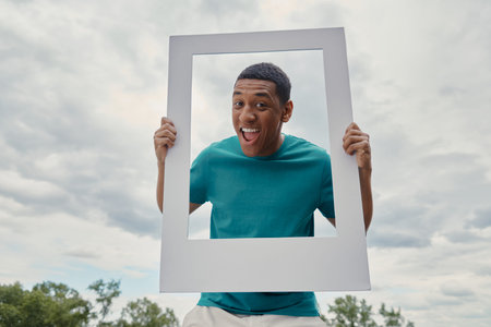 Cheerful mixed race man looking through a picture frame while standing outdoorsの写真素材