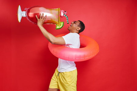 Playful young man carrying inflatable ring and cocktail while standing against red background