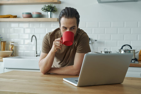 Handsome young man using laptop and enjoying coffee while leaning at the kitchen islandの写真素材
