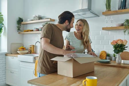 Beautiful young couple unpacking box and smiling while standing at the domestic kitchenの写真素材