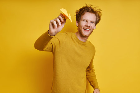 Happy redhead man holding paper airplane and smiling while standing against yellow background