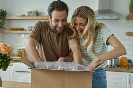 Happy young couple unpacking box while standing at the domestic kitchenの写真素材