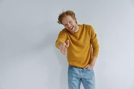 Cheerful redhead man stretching out hand for shake and smiling while standing against wallの写真素材