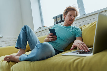 Handsome redhead man using smart using technologies while sitting on the couch at home