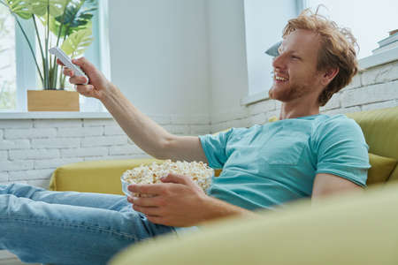 Handsome young man watching TV and enjoying snacks while sitting on the couch at homeの写真素材