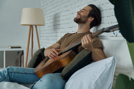 Happy young man playing acoustic guitar while relaxing on bed at home