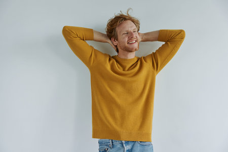 Relaxed young redhead man holding hands behind head while standing against wallの写真素材