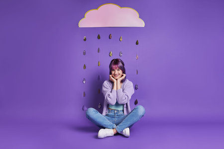 Cheerful young woman sitting under the paper cloud and smiling with purple background