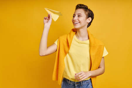 Happy young woman holding paper airplane while standing against yellow backgroundの写真素材