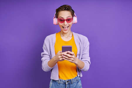 Excited woman in headphones looking at smart phone while standing against purple backgroundの写真素材