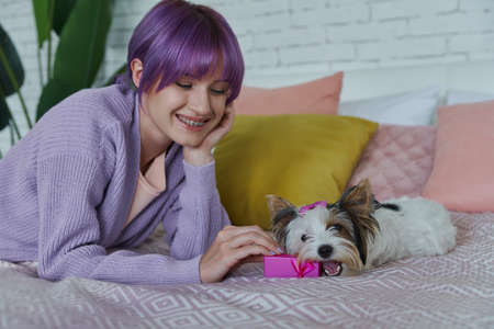 Playful young woman holding little gift box while lying in bed with her dogの写真素材