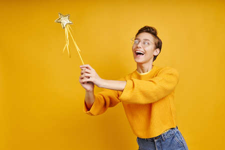 Playful young woman holding magic wand while standing against yellow backgroundの写真素材