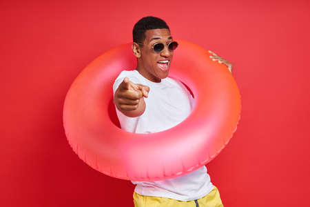 Playful young man carrying inflatable ring and gesturing while standing against red backgroundの写真素材