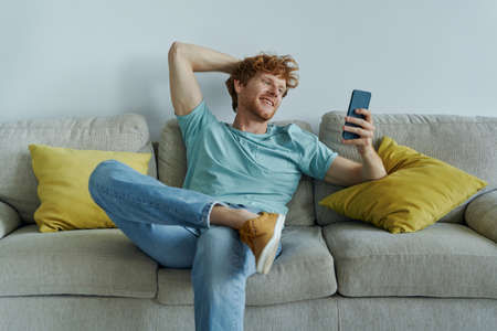 Handsome redhead man using smart phone while relaxing on the couch at homeの写真素材