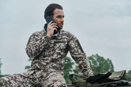 Confident young man in military uniform talking on mobile phone while sitting outdoorsの写真素材