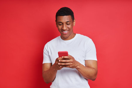Cheerful African man using smart phone while standing against red background
