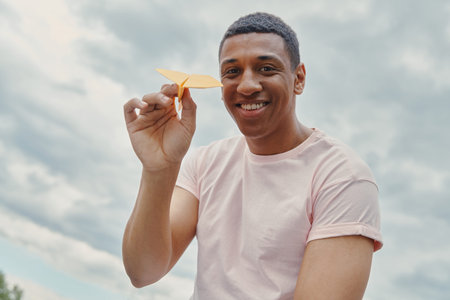 Low angle view of cheerful African man holding paper airplane with sky in the background