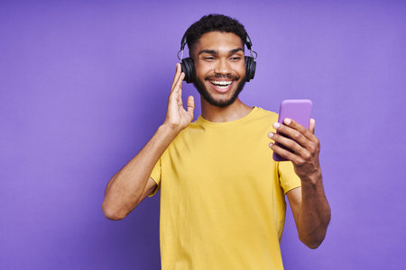 Handsome African man in headphones holding smart phone while listening music against purple...