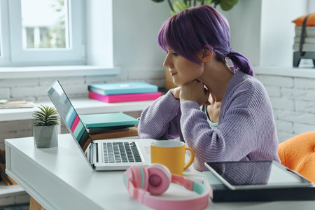 Concentrated young woman with purple hair working on laptop while sitting at her working place