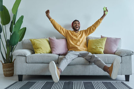 Happy young African man holding smart phone and gesturing while sitting on the couch