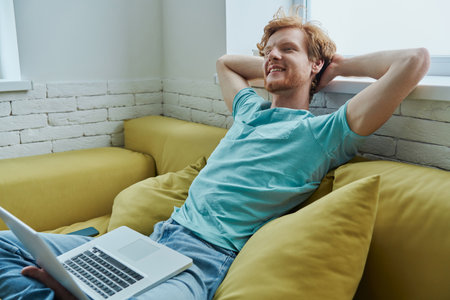 Cheerful redhead man using laptop and holding hands behind head while sitting on the couchの写真素材