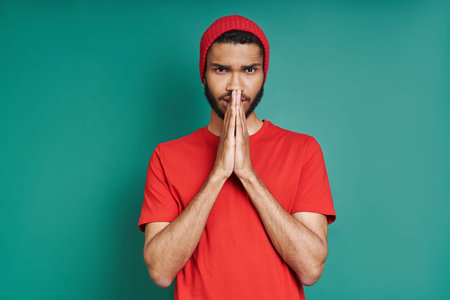 Worried African man holding hands clasped while standing against green background