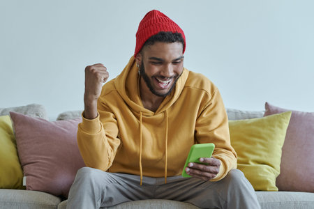 Excited young African man using smart phone and gesturing while sitting on the couch