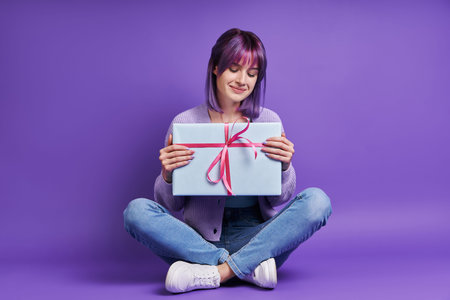 Beautiful young woman holding gift box and smiling while sitting against purple background