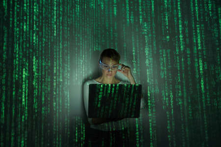 Confident woman adjusting futuristic glasses while using laptop against dark backgroundの写真素材