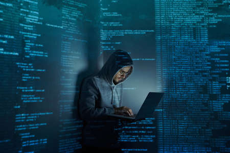Young man in hooded shirt using computer while standing against dark backgroundの写真素材