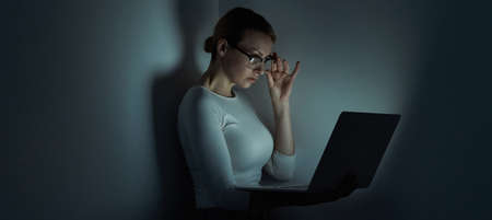 Beautiful young woman adjusting her glasses while working on laptop against a dark wallの写真素材