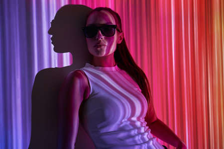 Beautiful young woman in eyeglasses leaning at the wall with colorful lightingの写真素材