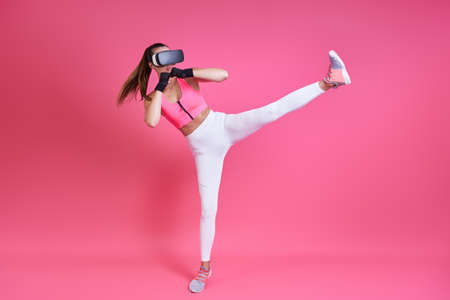 Young woman in virtual reality glasses kicking against pink backgroundの写真素材