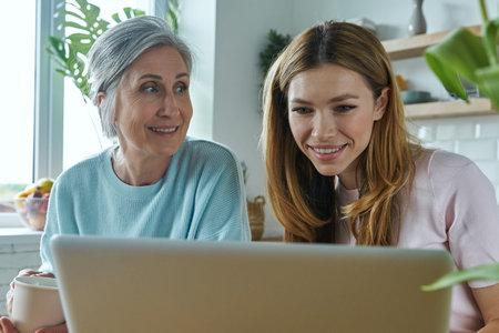 Happy mother and her adult daughter using laptop while spending time at home together