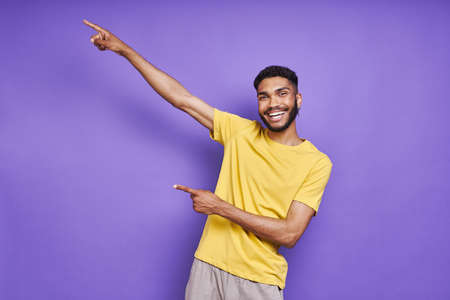 Handsome African man pointing away and smiling while standing against purple backgroundの写真素材