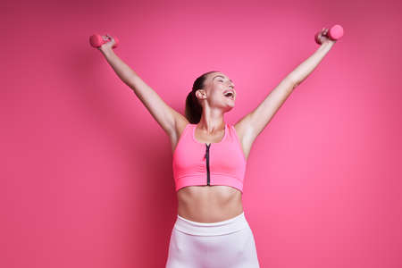 Happy young woman in sports clothing exercising with dumbbells against pink backgroundの写真素材