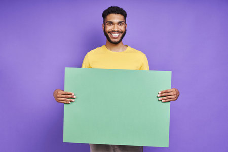 Happy African man holding green banner while standing against purple backgroundの写真素材