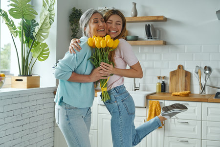 Happy senior mother receiving a bunch of tulips from her daughter while standing at the kitchen