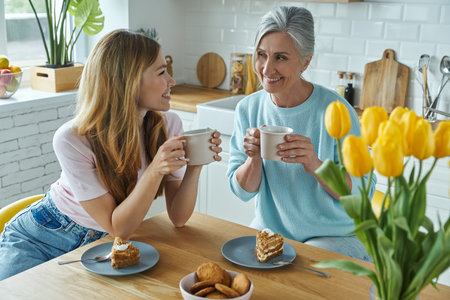 Senior mother and her adult daughter enjoying hot drinks and sweet food at the kitchenの写真素材