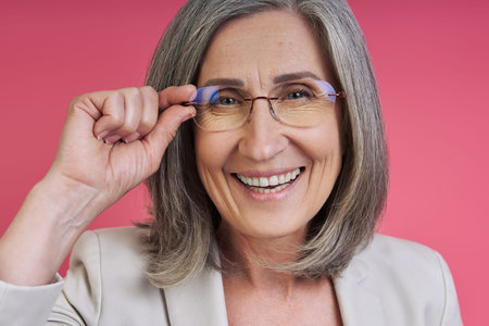 Happy senior woman in formalwear adjusting eyeglasses while standing against pink backgroundの写真素材