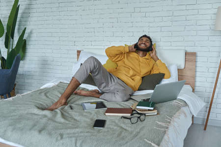 Happy young African man in headphones enjoying music while lying in bed at homeの写真素材