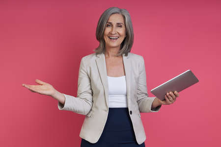 Cheerful senior woman in formalwear holding digital tablet while standing against pink backgroundの写真素材