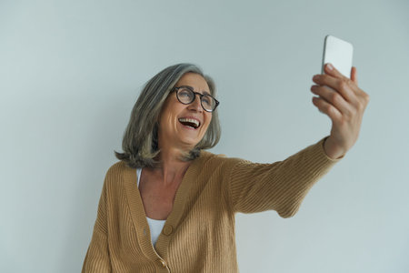 Happy senior woman making selfie by her smart phone while standing against white wall